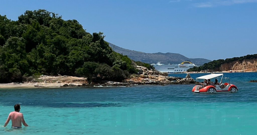 Strand von Ksamil – Ort mit guter Wasserqualität. Strandszene mit Tretboot, Badendem und im Hintergrund vor den Hängen von Korfu einer grossen Fähre